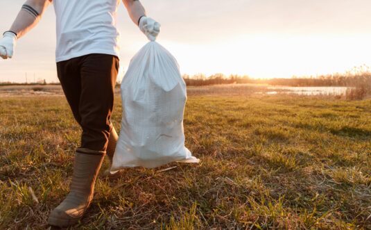 A volunteer carrying a garbage bag during a clean-up in a sunny outdoor field.