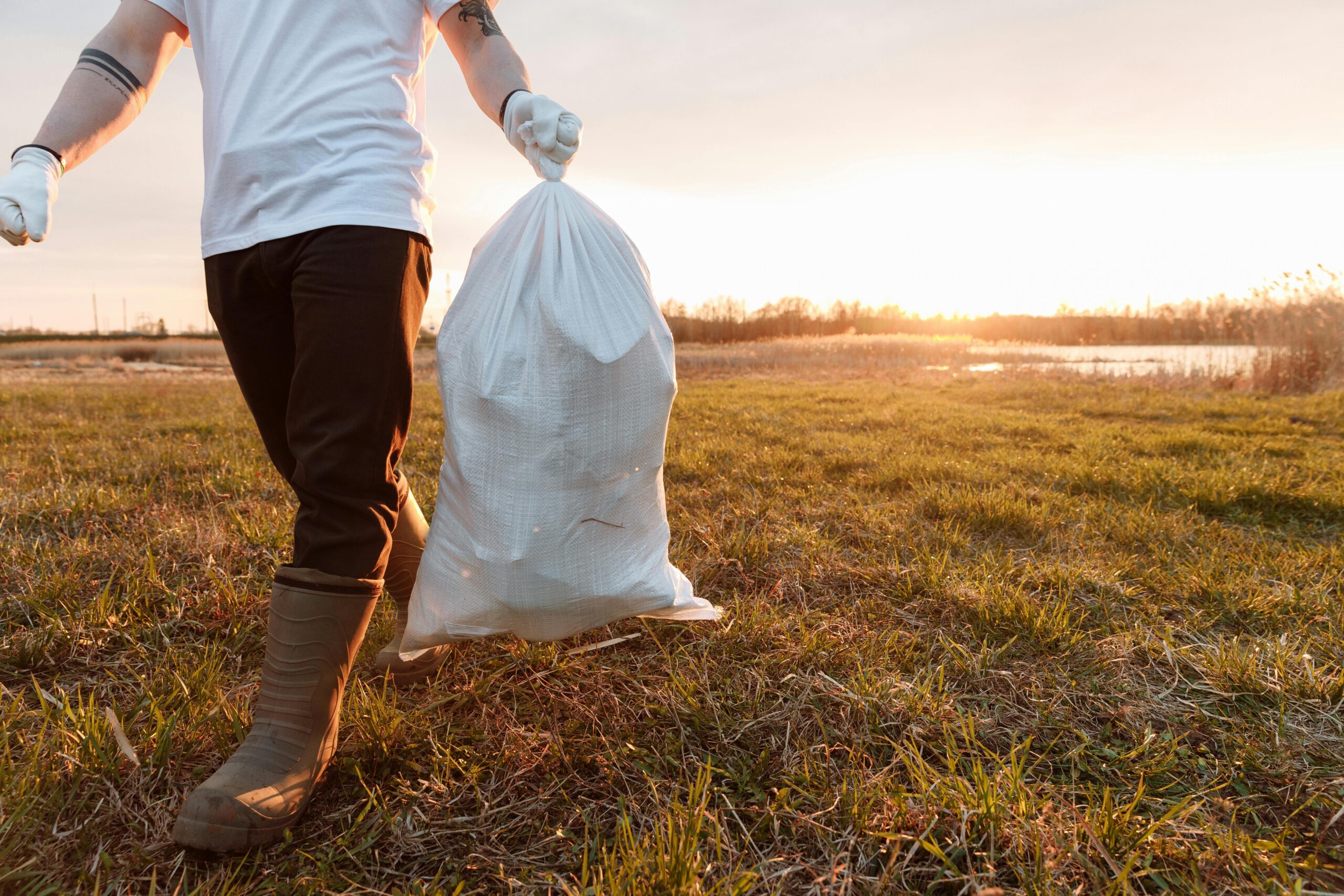 A volunteer carrying a garbage bag during a clean-up in a sunny outdoor field.